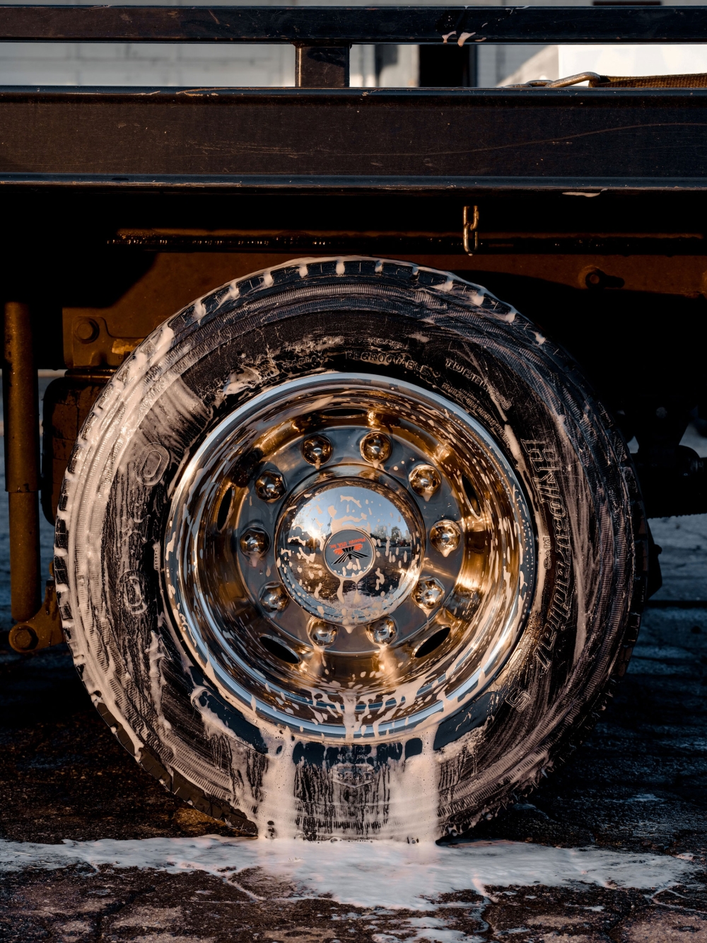 Close-up shot of a truck tire with soap suds running down the sidewall, set against a black truck body and a paved surface.