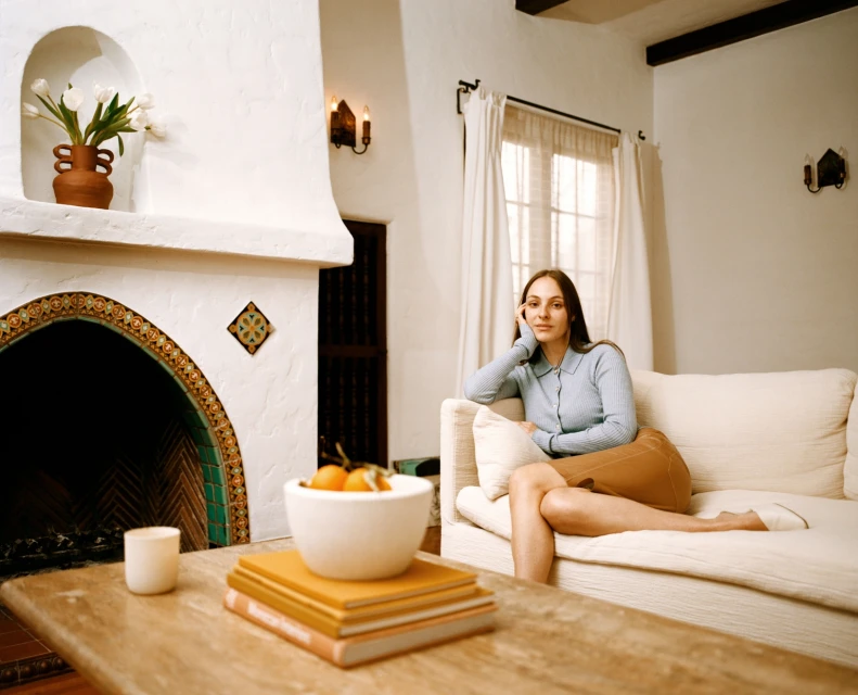 Indoor portrait of a woman with long brown hair sitting on a white sofa with a tan skirt, blue top, in front of a fireplace.