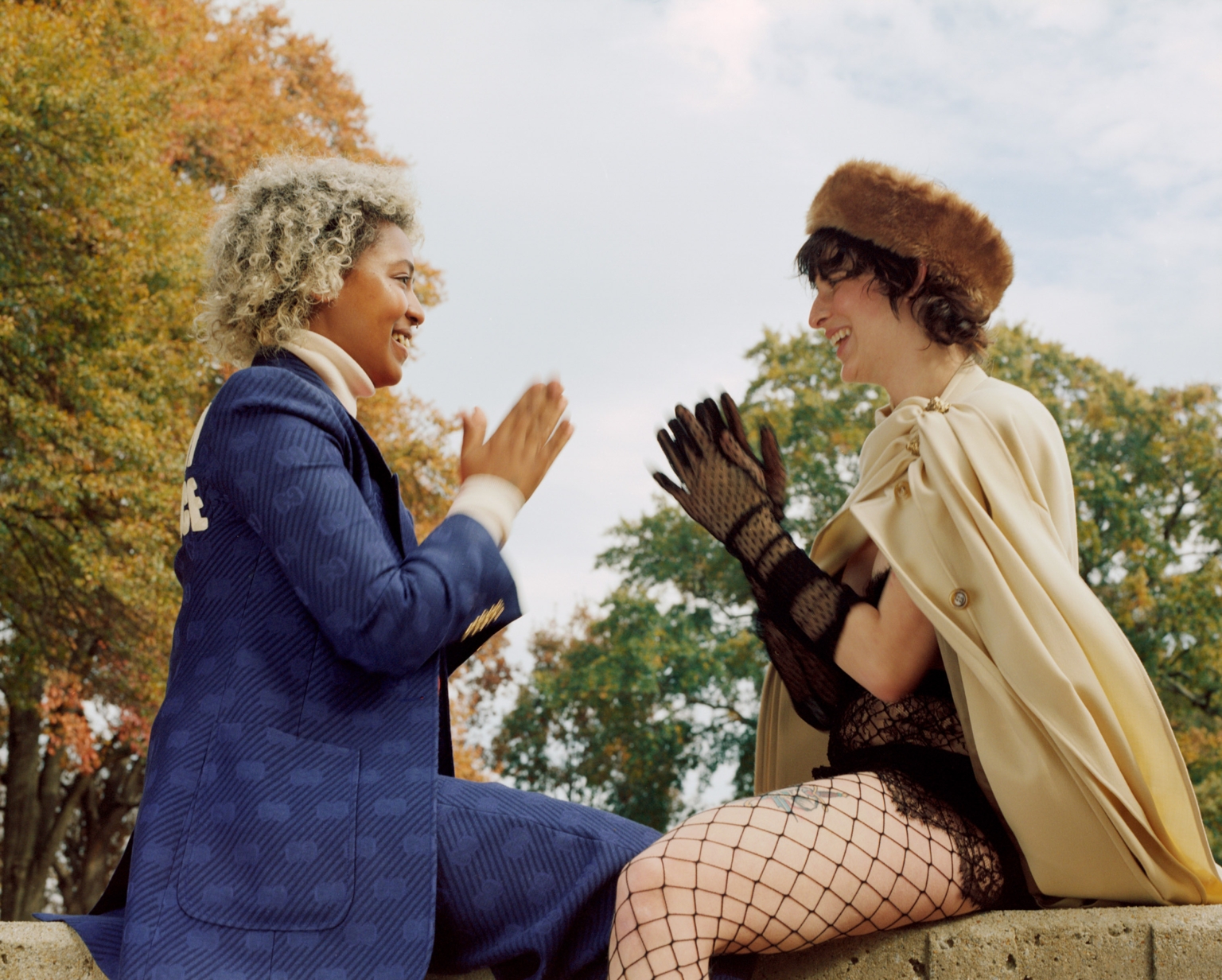 Two women in fashionable vintage attire sitting and chatting outside in a park during the day, surrounded by autumn trees and a blue sky.