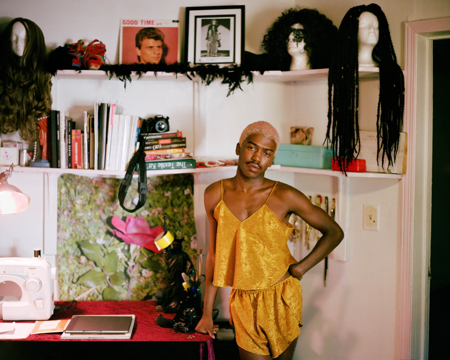 Young adult posing in a golden, patterned, silk pajama set in an indoor, home-like setting with shelves and decorative objects.
