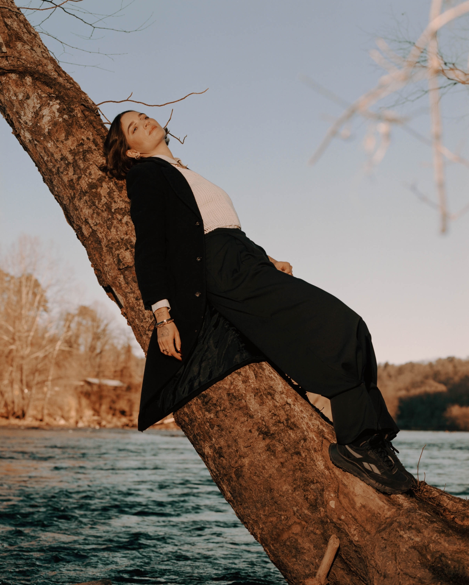 Woman in black coat leans against a tree near a river under a blue sky; portrait-oriented lifestyle fashion shot in an outdoor environment.