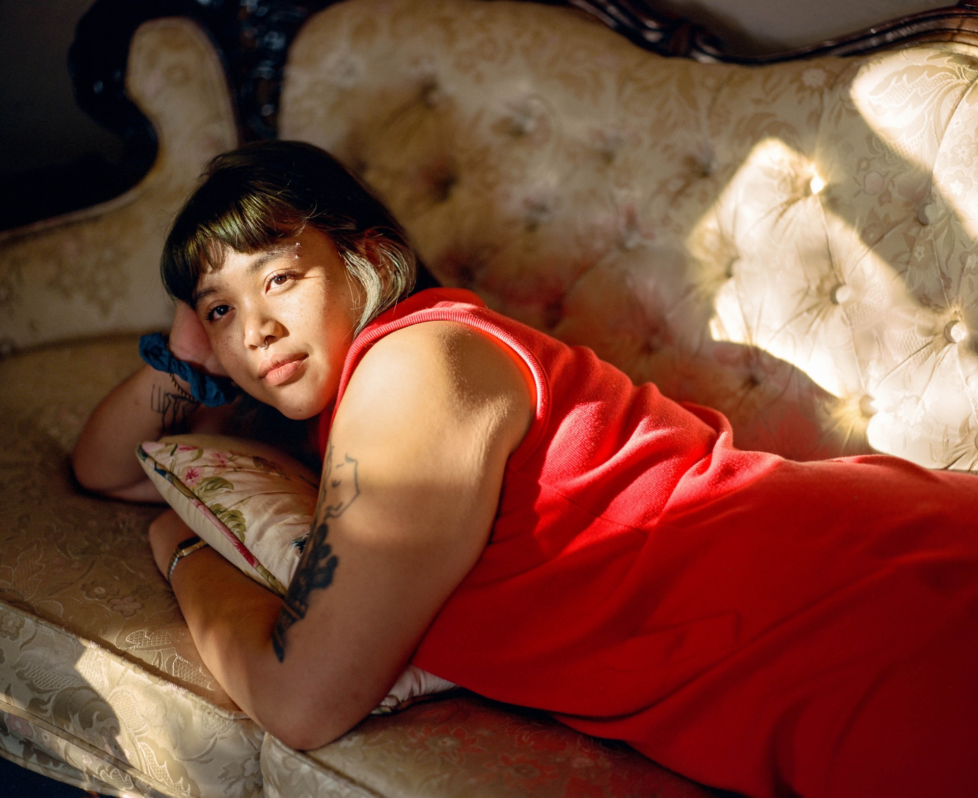 Portrait of an adult woman with dark hair in a red dress, lying on an ornate white couch in a sunlit room. Head and shoulders.