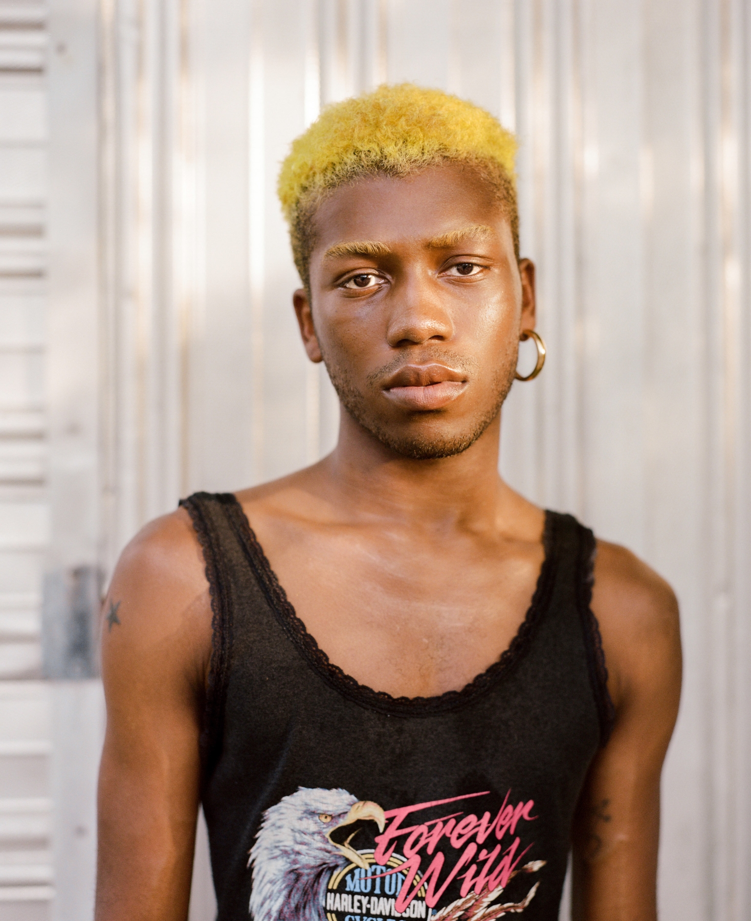 Close-up portrait of a young adult with blond hair, wearing a graphic tank top, and gold hoop earring against a white wall.