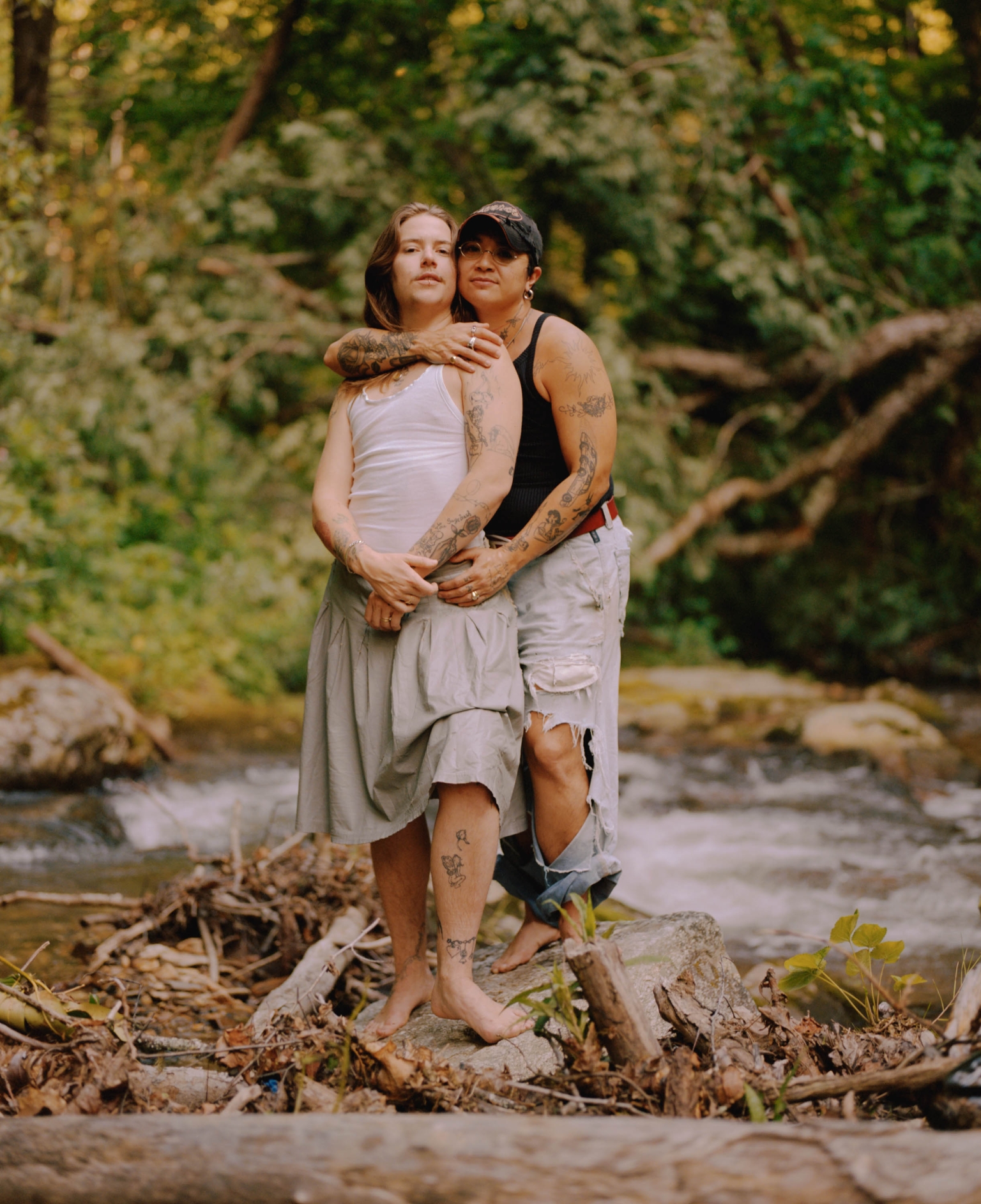 Two people stand barefoot, hugging near a stream, surrounded by lush green trees and foliage, with soft, natural light filtering through.