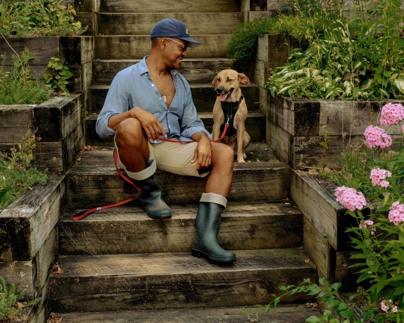 A man sits on wooden stairs with his dog, surrounded by lush greenery and pink flowers, enjoying a peaceful moment outdoors.