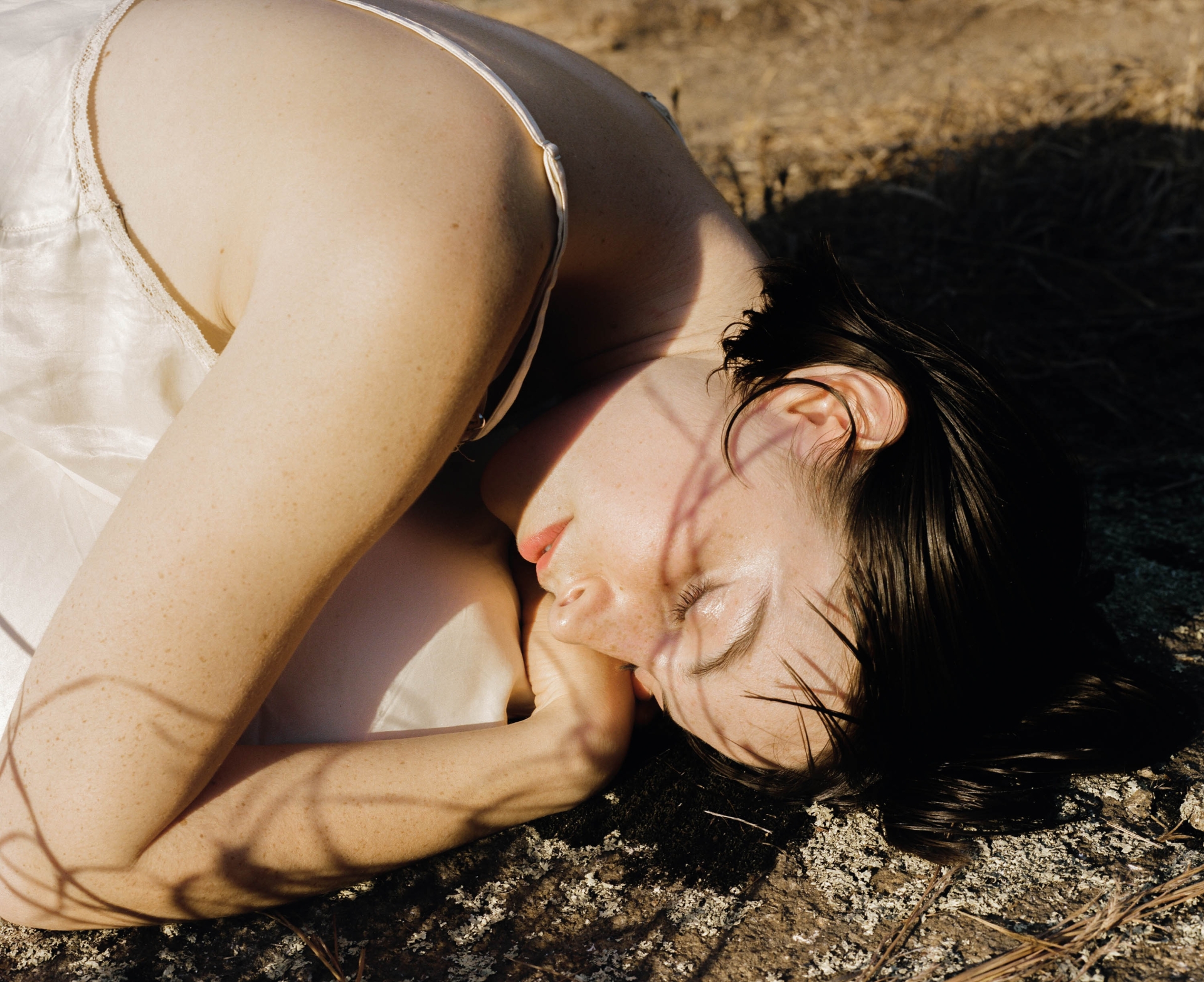 Close-up of a woman lying down outdoors in a dress, her face tilted to the side, with soft lighting and shadows from tree branches.
