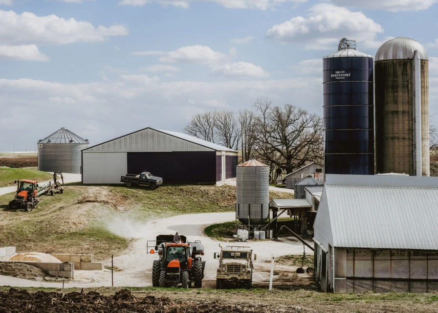 Rural farmland scene with tractors, farm trucks, barns, and grain silos under a partly cloudy sky, capturing the essence of agriculture.