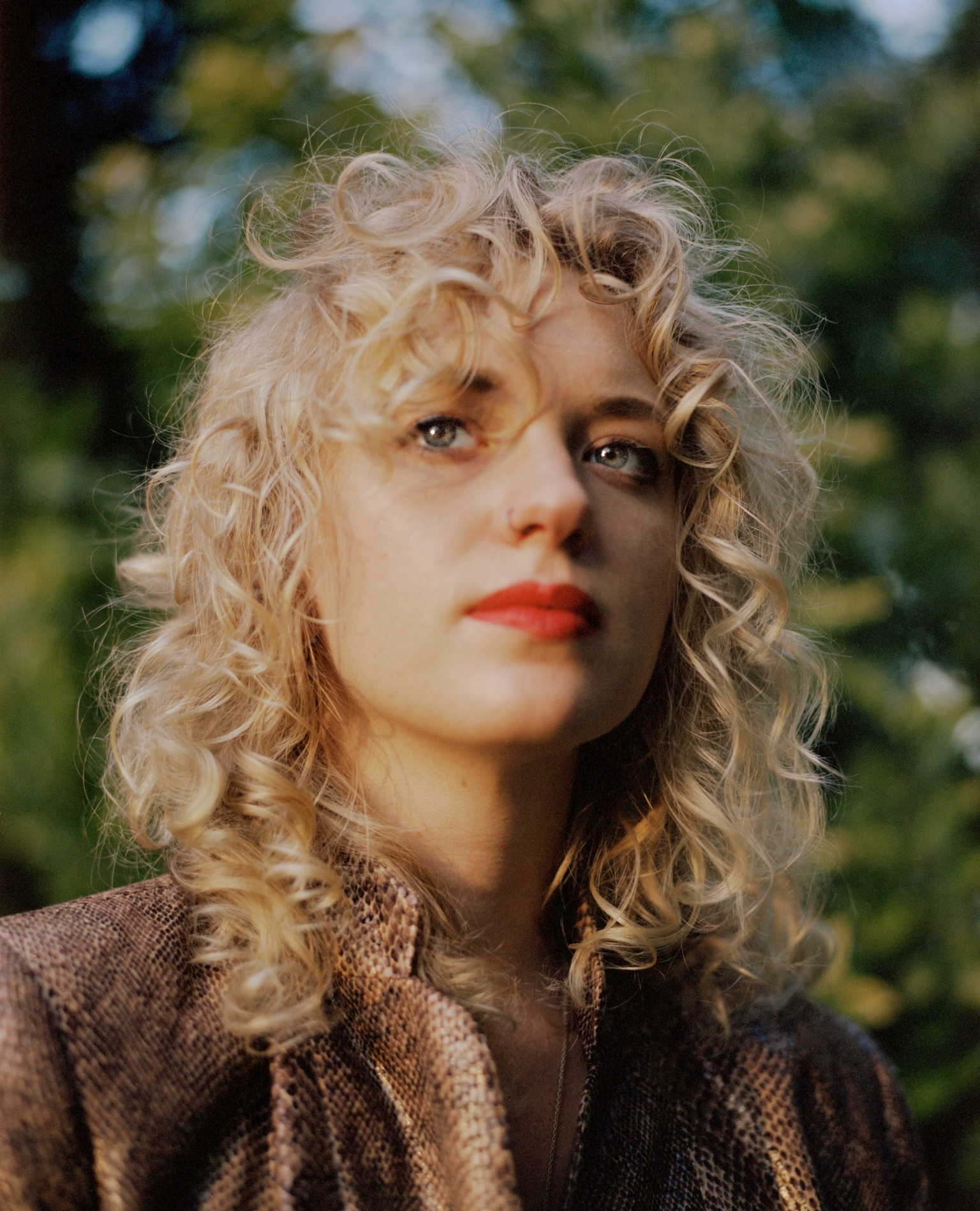 Close-up portrait of a young adult woman with curly blonde hair and red lipstick, looking up slightly, against a blurred green background.