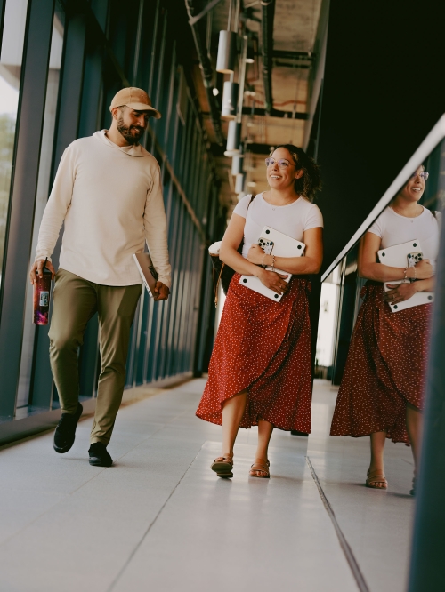 Two people walking in a hallway; a man in a cream hooded sweatshirt and woman in a red skirt carry a laptop, with the woman's reflection.