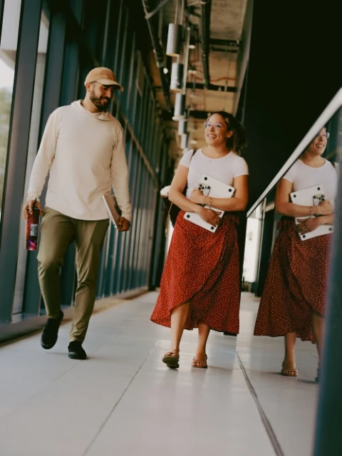 Two people walking in a hallway; a man in a cream hooded sweatshirt and woman in a red skirt carry a laptop, with the woman's reflection.