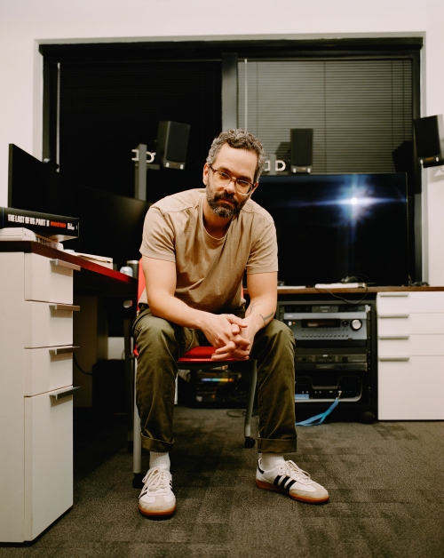 Portrait of a man with a beard wearing glasses and casual clothes, sitting on a red chair in an indoor office environment with computer equipment.