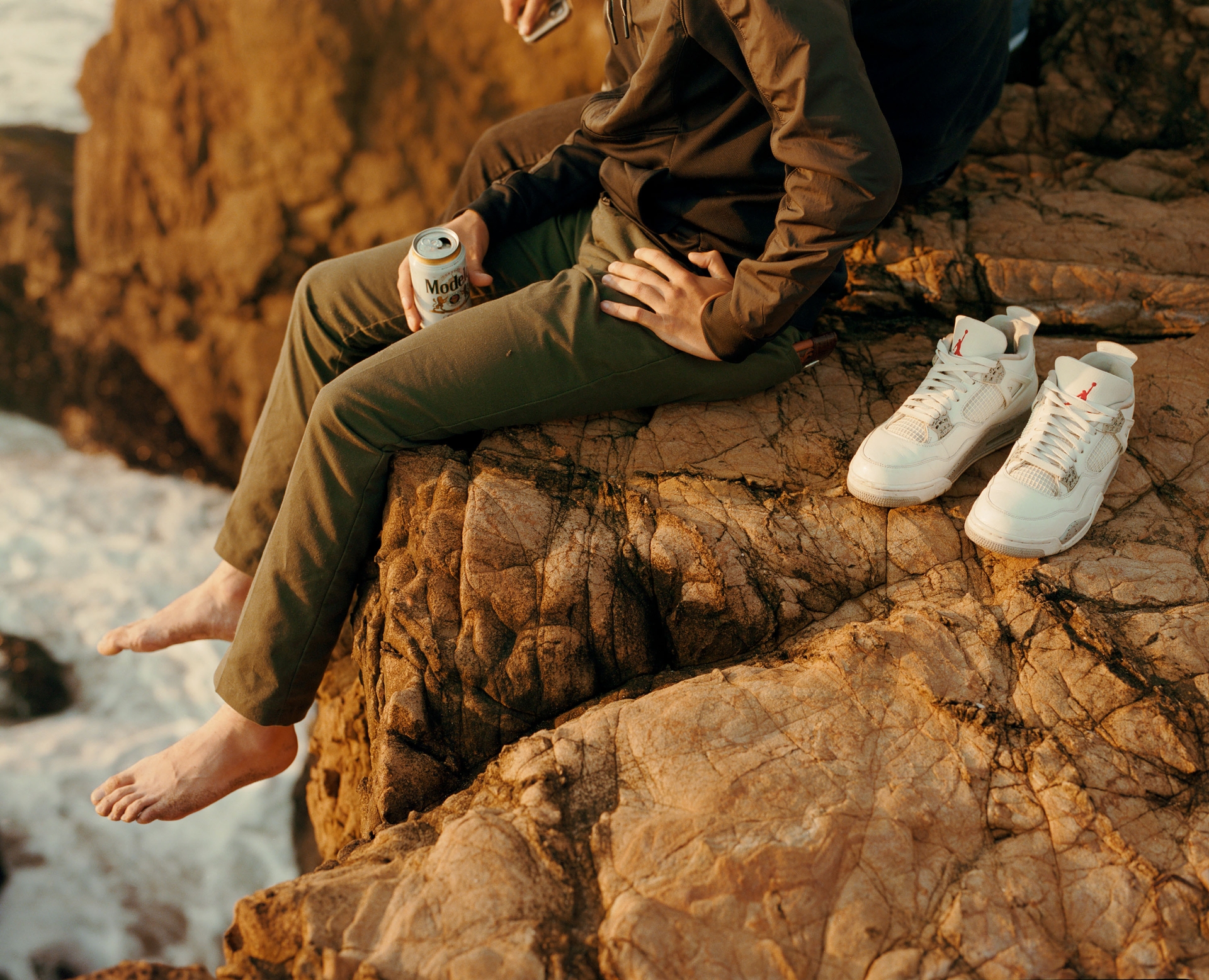 Two men sit on a rocky cliff overlooking the ocean, showcasing white sneakers and a Modelo beer in warm, natural light during daytime.