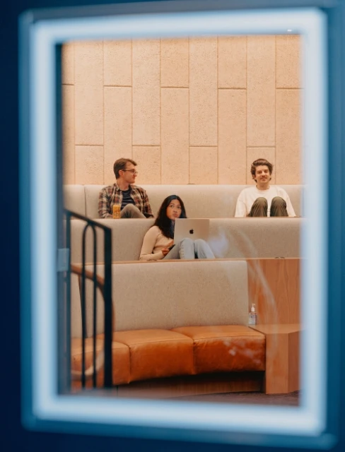 Three people sit on a tiered seating arrangement in a brightly lit indoor space, framed by a window with a dark blue frame.