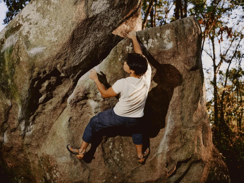 Adult male rock climbing on a large boulder in a forest. He is wearing a white t-shirt, dark pants, and climbing shoes.