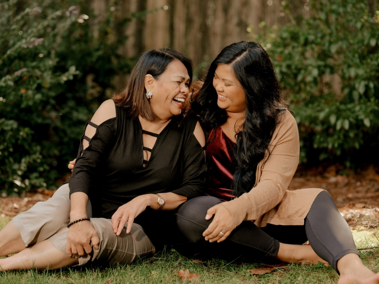 Two smiling women sit together on green grass near a wooded area, one in black and one in burgundy and tan.