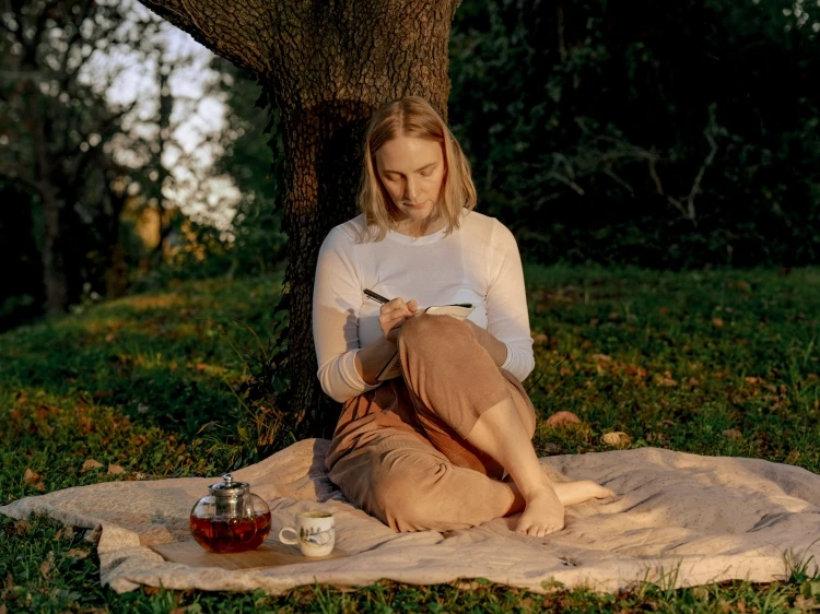 Young woman sitting under a tree writing in a journal outdoors, with a teapot and teacup on a blanket on the grass.