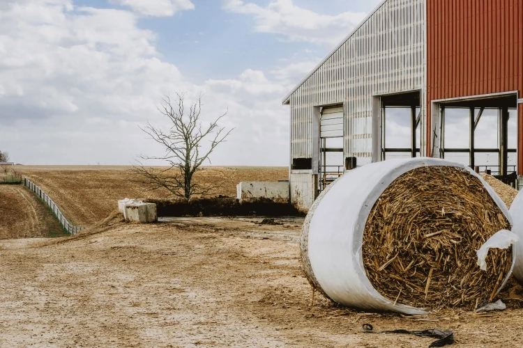 Rural landscape features a large hay bale in the foreground, with a barn and farm field visible in the background under a cloudy sky.