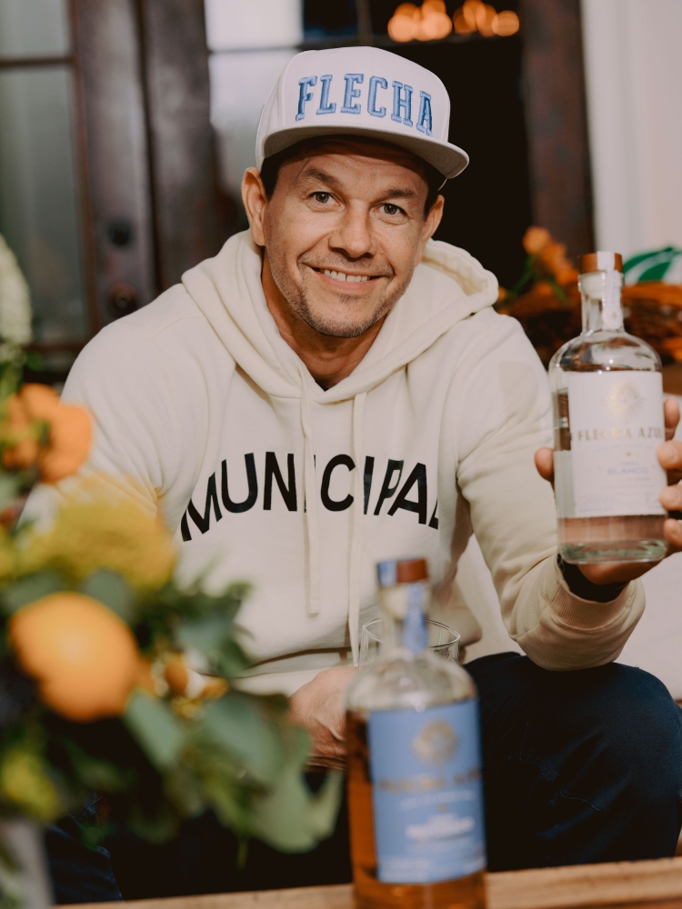 Adult male in a sweatshirt and baseball cap holds two liquor bottles in an indoor setting, with a vase of orange flowers visible.
