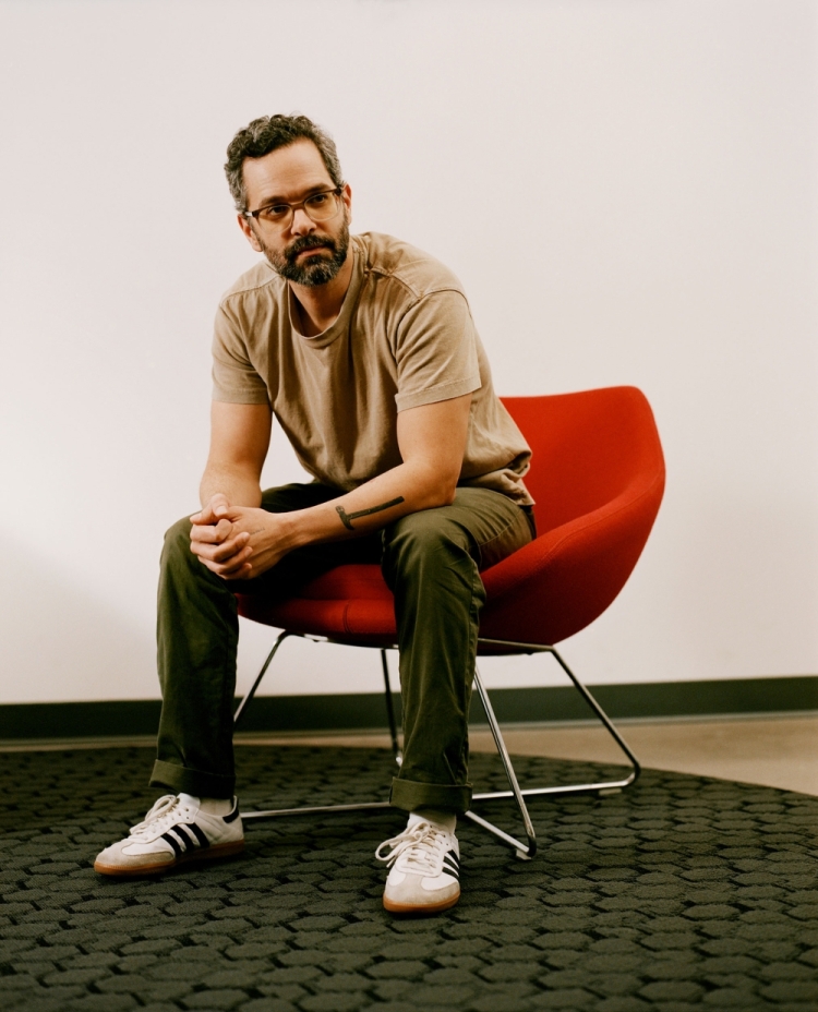 Bearded man in a taupe t-shirt sits in a red chair, casually posed in front of a white wall with a gray patterned carpet.