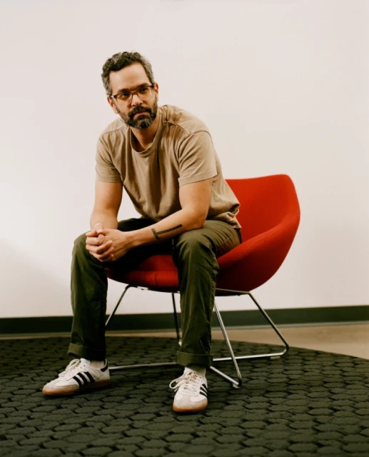 Bearded man in a taupe t-shirt sits in a red chair, casually posed in front of a white wall with a gray patterned carpet.
