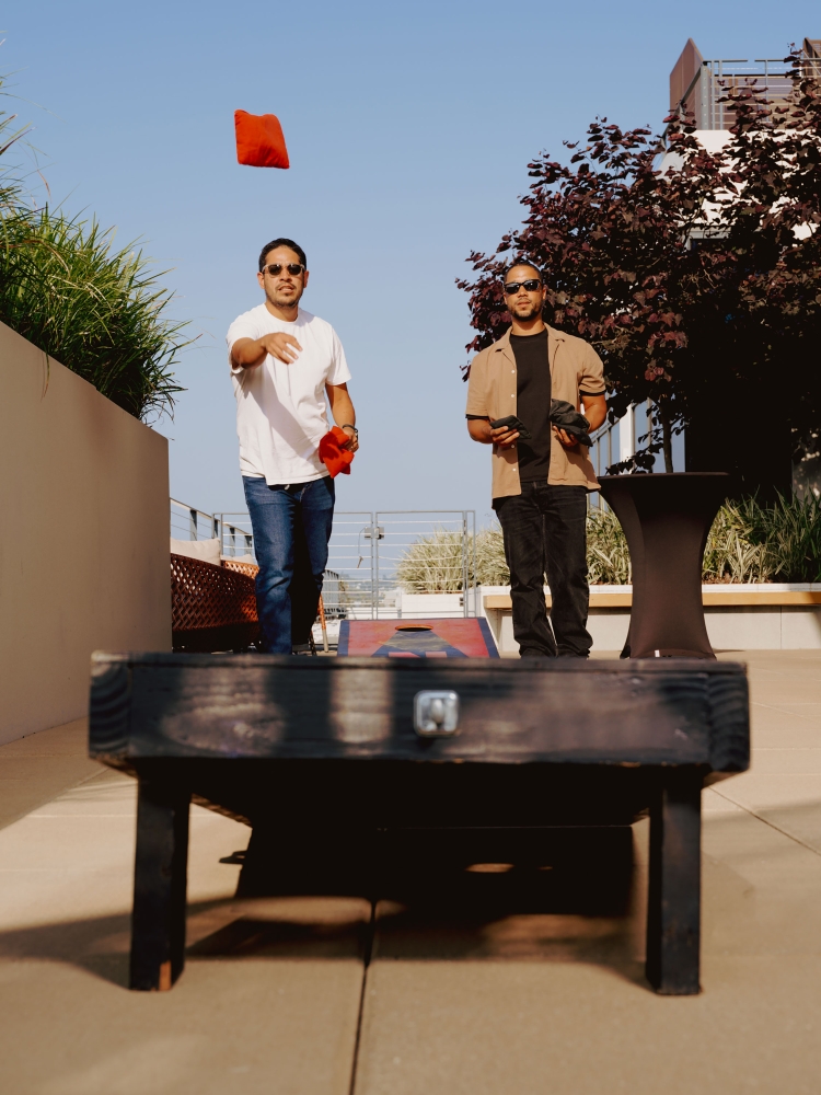 Two adult men play cornhole outdoors on a sunny day, one throwing a red beanbag toward the cornhole platform.