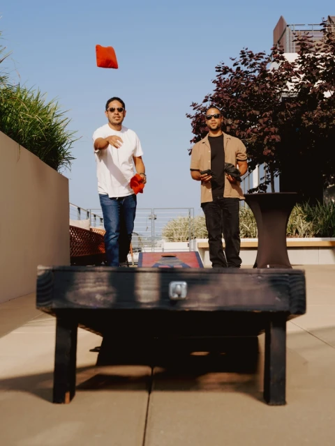 Two adult men play cornhole outdoors on a sunny day, one throwing a red beanbag toward the cornhole platform.