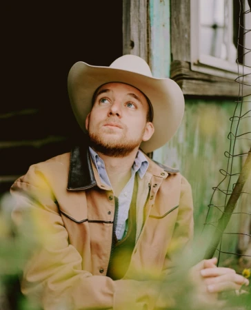 A man wearing a cowboy hat and tan jacket looking upward in a rustic outdoor setting photographed by Peyton Fulford.