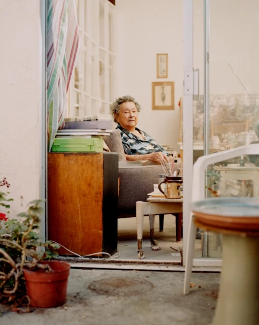 Elderly woman with short curly grey hair sitting on a grey couch in a brightly lit room with a doorway to an outdoor patio.