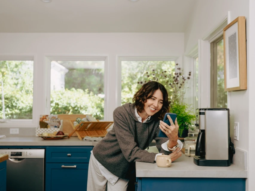 Woman smiles while looking at her phone in a naturally lit kitchen. The kitchen has blue cabinets, a stainless steel dishwasher, and a coffee maker.