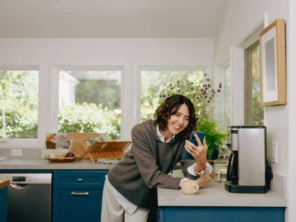 Woman smiles while looking at her phone in a naturally lit kitchen. The kitchen has blue cabinets, a stainless steel dishwasher, and a coffee maker.