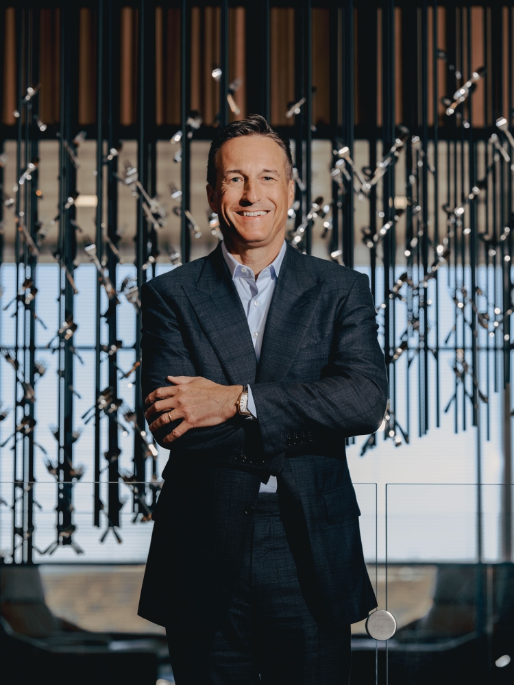 Portrait of a mature adult man in a suit standing indoors with arms crossed in front of a modern architectural decorative background.