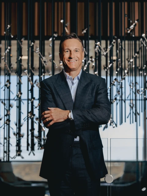 Portrait of a mature adult man in a suit standing indoors with arms crossed in front of a modern architectural decorative background.