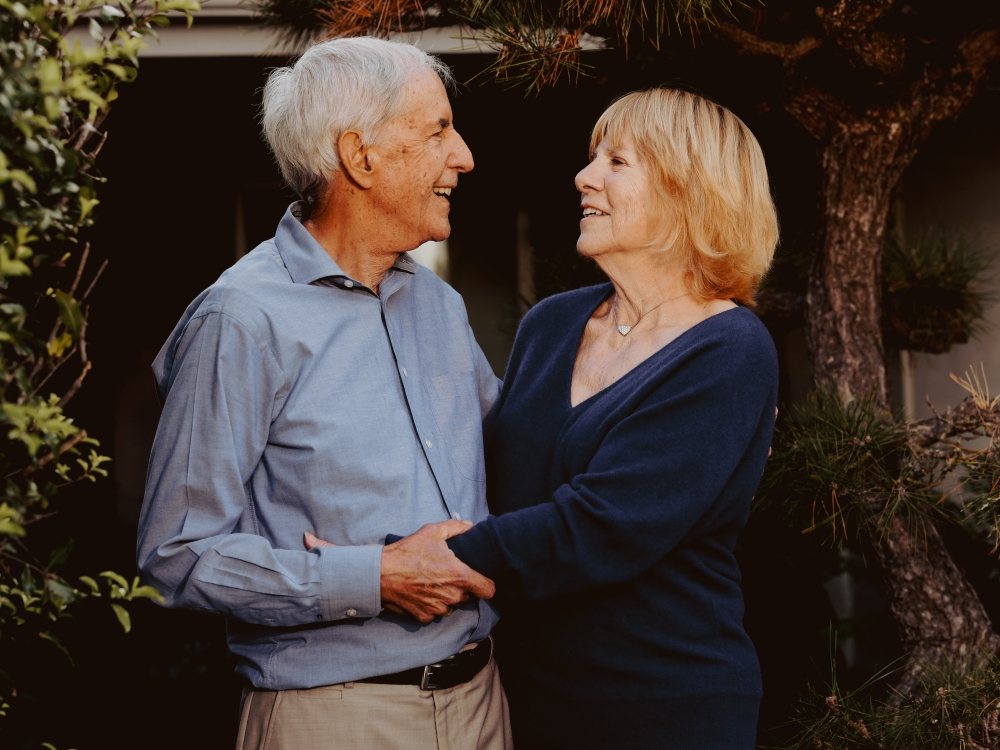 Elderly couple embraces outdoors, smiling at each other in a medium shot with natural light, wearing blue shirts and standing near greenery.