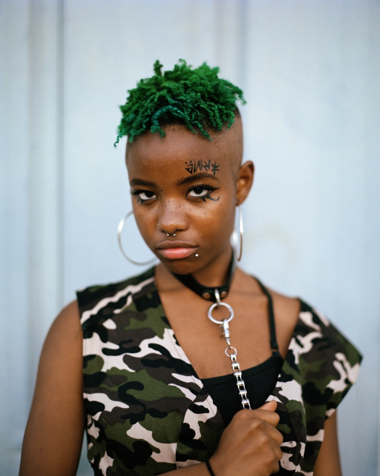 A young adult female with vibrant green hair, facial piercings, and a camouflage vest poses indoors with a neutral facial expression.