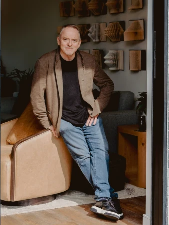 Middle-aged man wearing a brown blazer, black t-shirt, and blue jeans leaning against a leather armchair inside a living room
