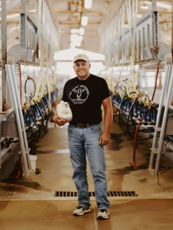 Mature man in t-shirt and cap holding milk jug in a commercial dairy barn with milking equipment. Indoors in agricultural setting.