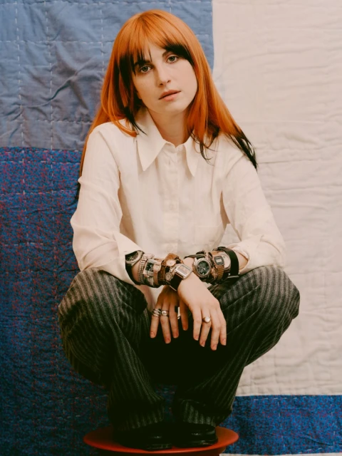 Red-haired adult female poses indoors wearing white blouse, pinstripe pants, many wrist watches, and rings in a studio setting.