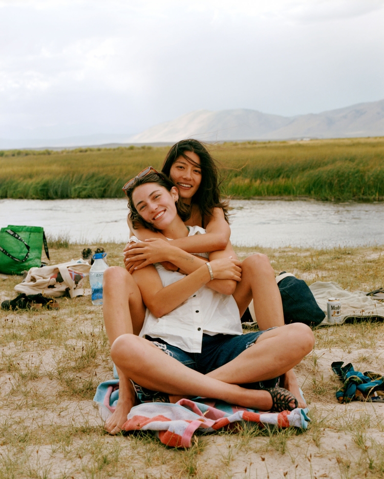 Two women embrace on a sunny beach near a river with distant mountains, creating a heartwarming scene of friendship and natural beauty.