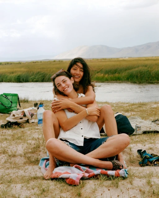 Two women embrace on a sunny beach near a river with distant mountains, creating a heartwarming scene of friendship and natural beauty.