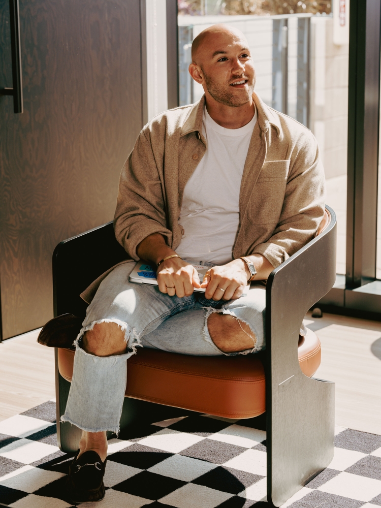 Stylish man with short hair sits in a modern chair inside a bright interior wearing jeans, a white shirt, and a tan jacket.
