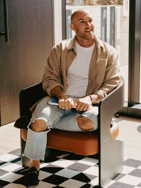 Stylish man with short hair sits in a modern chair inside a bright interior wearing jeans, a white shirt, and a tan jacket.