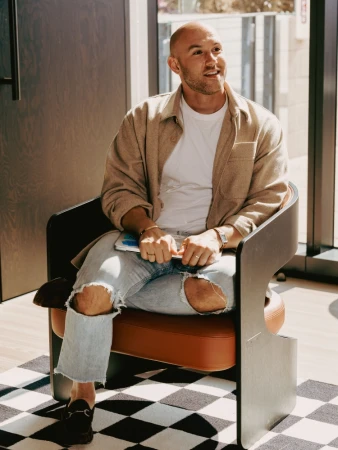 Stylish man with short hair sits in a modern chair inside a bright interior wearing jeans, a white shirt, and a tan jacket.