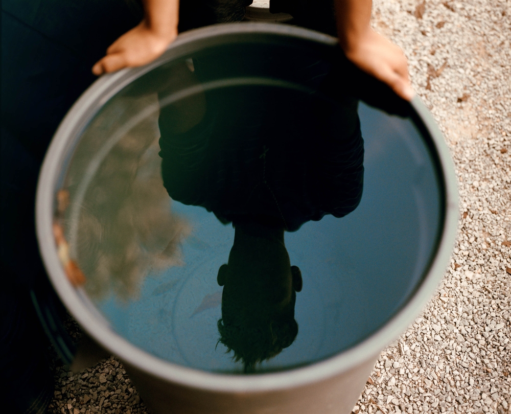 Child reflected in water-filled bucket. The water reflects the child's silhouette and the sky. Ground visible with small stones.