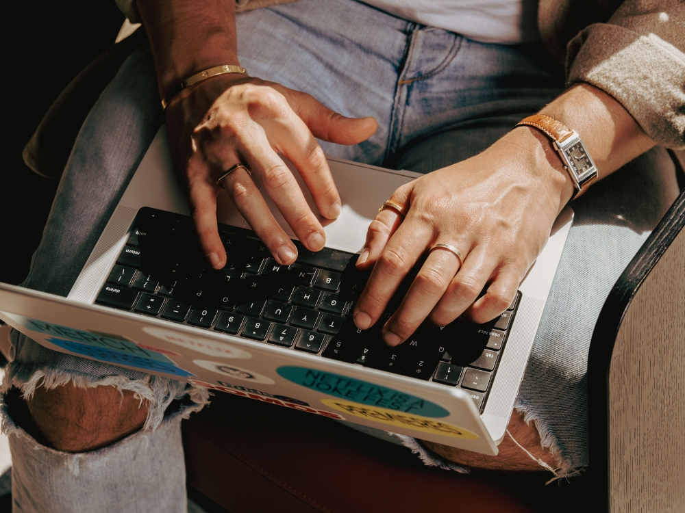 Close-up of hands typing on a laptop keyboard while wearing denim jeans, gold jewelry, and a wristwatch.