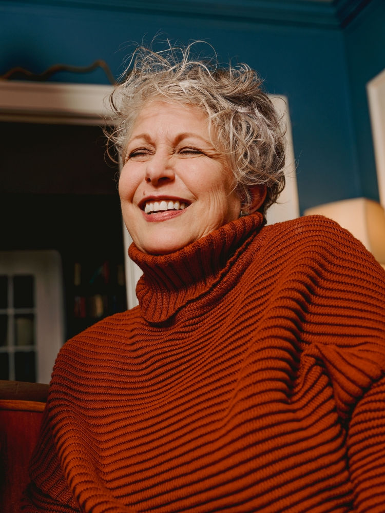 Smiling elderly woman with short gray hair wearing a rust-colored ribbed turtleneck sweater indoors with a teal wall in the background.
