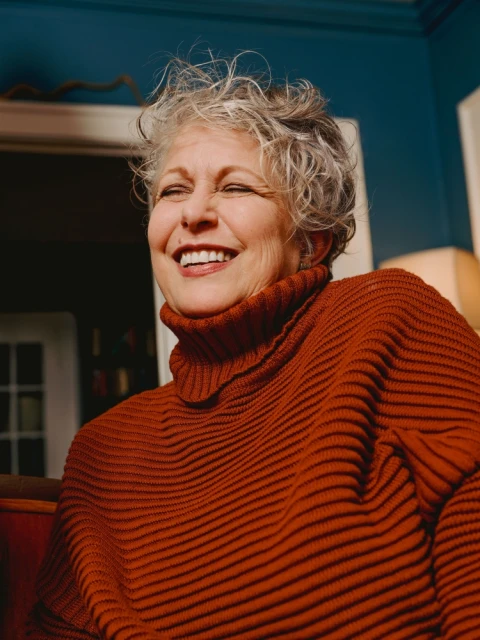 Smiling elderly woman with short gray hair wearing a rust-colored ribbed turtleneck sweater indoors with a teal wall in the background.