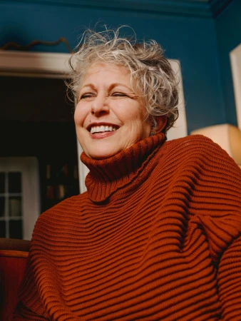 Smiling elderly woman with short gray hair wearing a rust-colored ribbed turtleneck sweater indoors with a teal wall in the background.