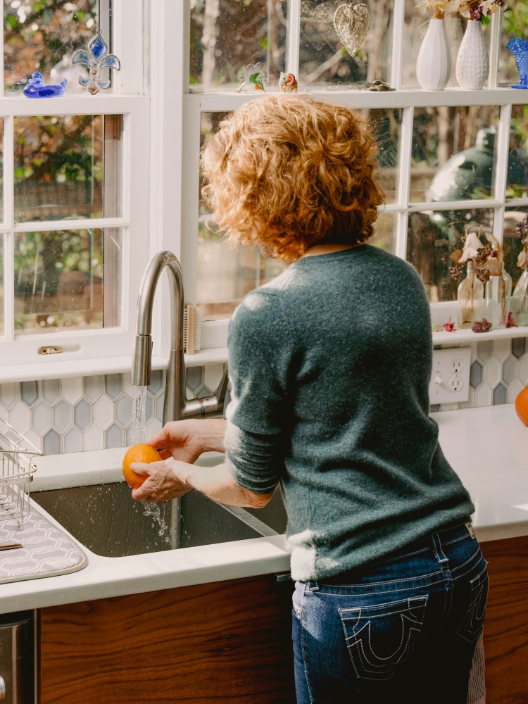 Woman washing an orange at a kitchen sink next to a window. The kitchen features white countertops and wood cabinets.