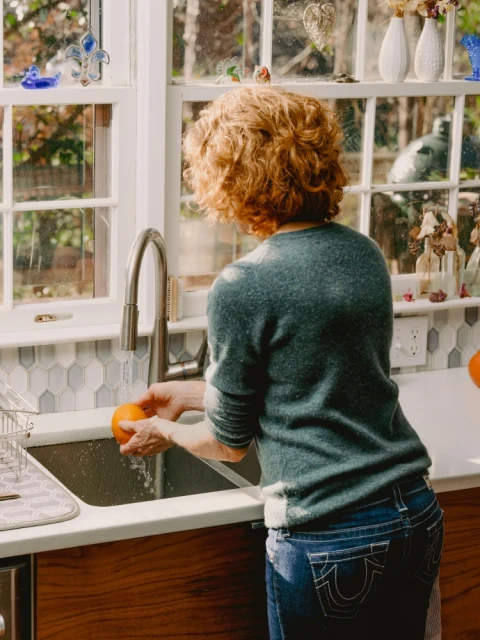 Woman washing an orange at a kitchen sink next to a window. The kitchen features white countertops and wood cabinets.