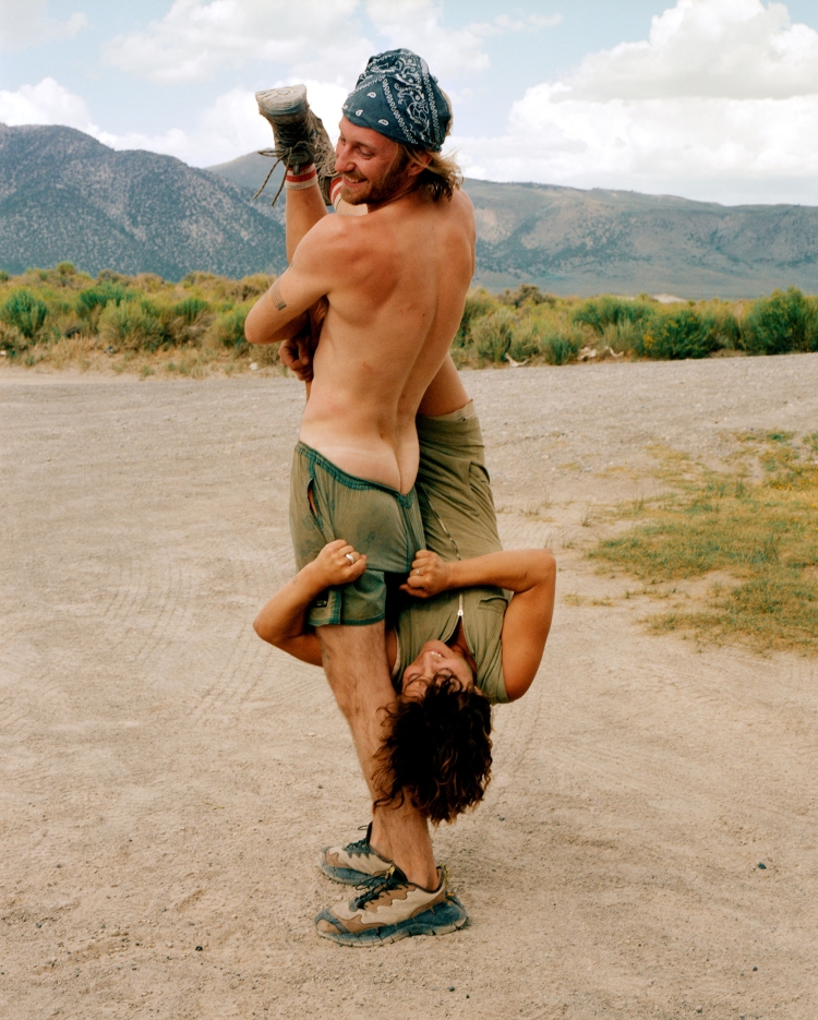 Man holding woman upside down in rural landscape. He's shirtless with shorts and a bandana; she wears a zip-up shirt and shorts.
